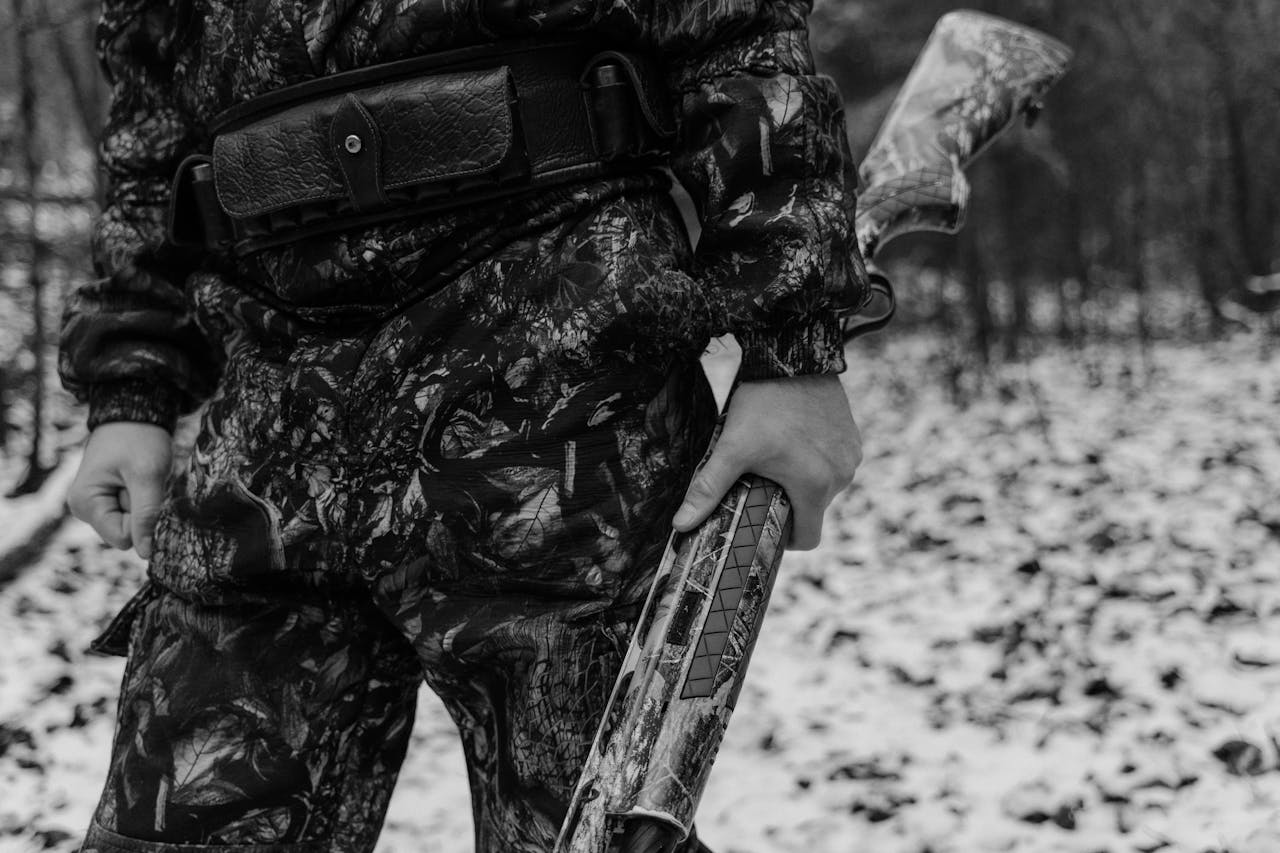 category-01 Monochrome image of a hunter in camouflage gear holding a shotgun in a snowy forest.